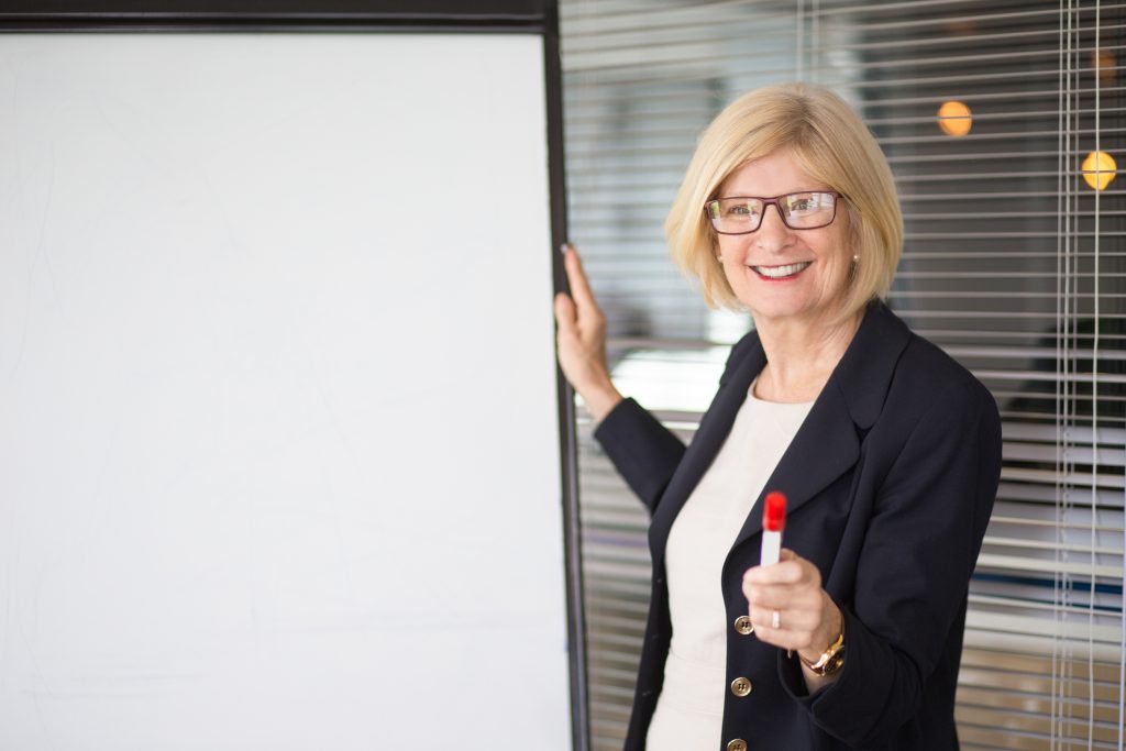 Closeup portrait of smiling attractive senior business woman looking at camera, giving lecture, addressing audience and standing at whiteboard with office partition in background